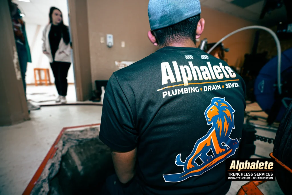 Alphalete trenchless plumbing technician inspecting an in-home floor excavation site while a homeowner stands in the background.