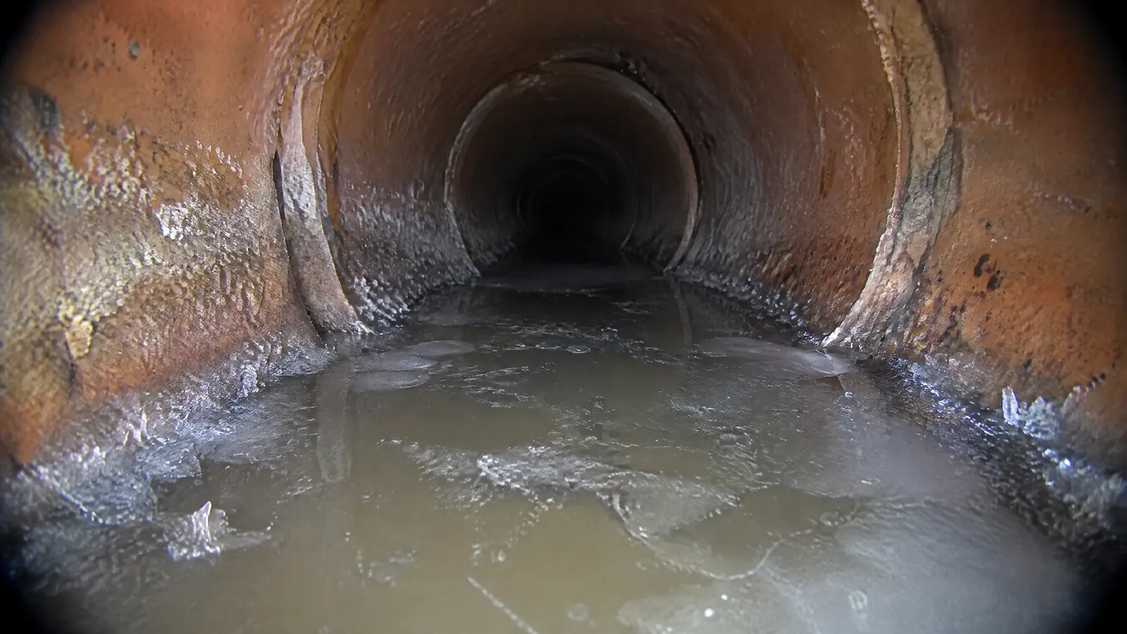 Sewer camera view inside a clay sewer pipe showing a sagging section holding standing water with a thin layer of ice